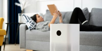 Closeup of an Air Purifier with a Woman Laying on Couch Reading a Book in the Background How to Improve Indoor Air Quality